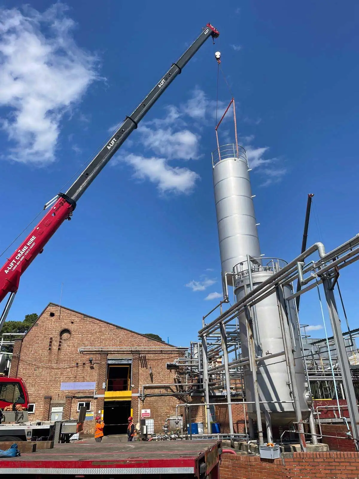A-Lift crane hoisting a metal cylinder at an industrial site under a blue sky