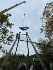 Crane lifting a metal platform above a tall A-frame structure among trees