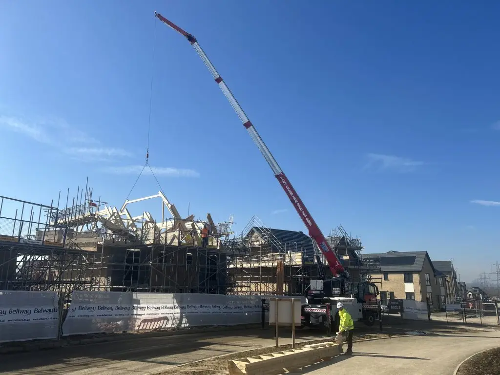 Mobile crane lifting a roof truss onto a scaffolded building under a clear blue sky