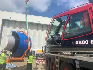 Crane truck lifting a large orange and blue cylinder beside workers in high-visibility vests