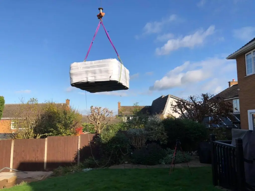 Wrapped pallet suspended by crane hook over a residential garden