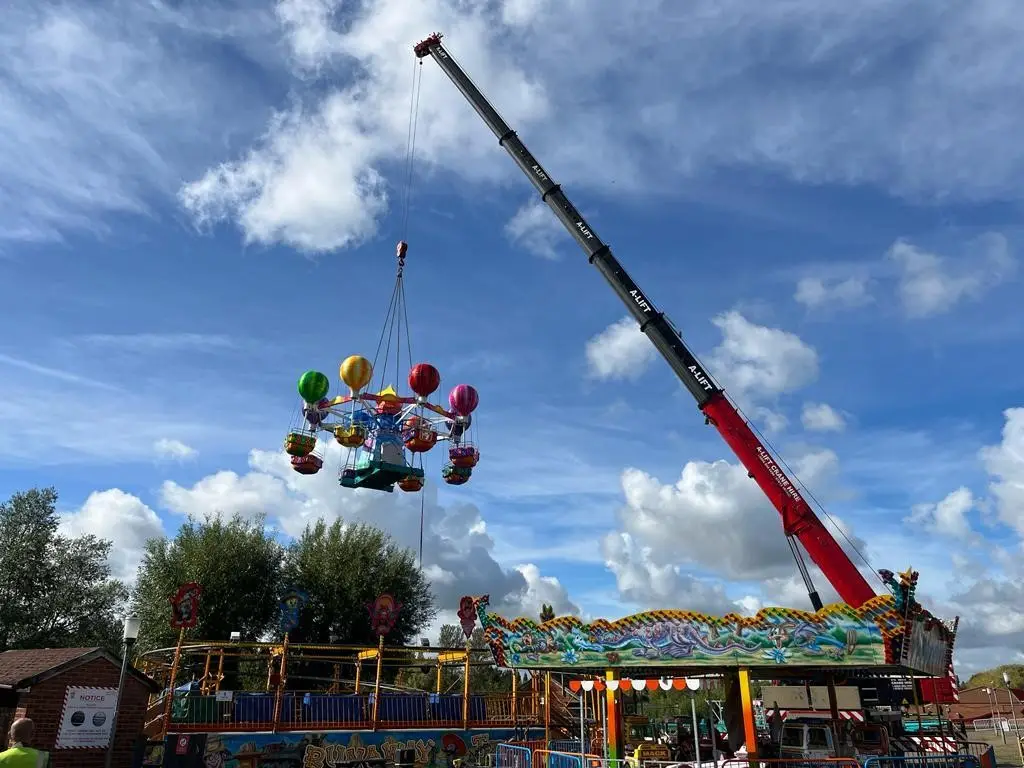 Crane lifting a colorful fairground ride under a blue, cloudy sky