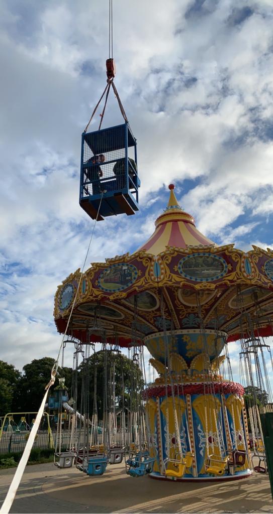 Crane lifting a blue cage above a colorful swing carousel under a cloudy sky